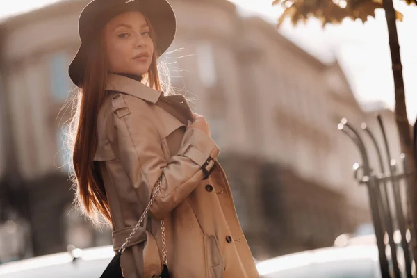 Стильна молода жінка в бежевому пальто в чорному капелюсі на міській вулиці. Жіноча вулична мода. Осінній одяг. Міський стиль — стокове фото