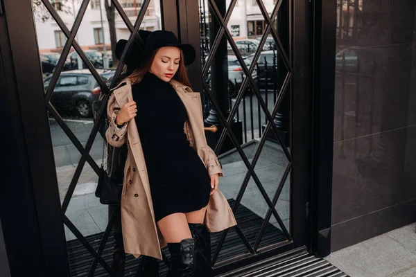 Stijlvolle jonge vrouw in een beige jas met een zwarte hoed op een straat. Vrouwenmode op straat. Herfst kleding. Stedelijke stijl — Stockfoto