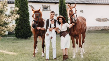 Beyaz elbiseli bir aile ve oğulları doğada iki güzel atın yanında dururlar. Çocuklu şık bir çift atlarla fotoğraf çekilir.