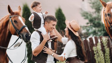 Beyaz elbiseli bir aile ve oğulları doğada iki güzel atın yanında dururlar. Çocuklu şık bir çift atlarla fotoğraf çekilir.