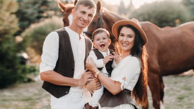 Beyaz elbiseli bir aile ve oğulları doğada iki güzel atın yanında dururlar. Çocuklu şık bir çift atlarla fotoğraf çekilir.