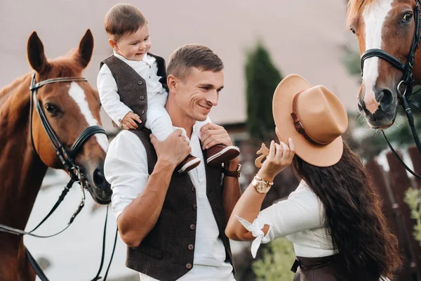Rodzina w białych ubraniach z synem stoi w pobliżu dwóch pięknych koni w przyrodzie. Stylowa para z dzieckiem jest fotografowana z końmi — Zdjęcie stockowe