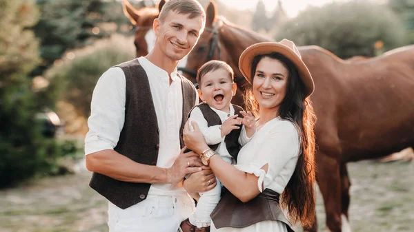 Una familia vestida de blanco con su hijo está cerca de dos hermosos caballos en la naturaleza. Una elegante pareja con un niño son fotografiados con caballos —  Fotos de Stock