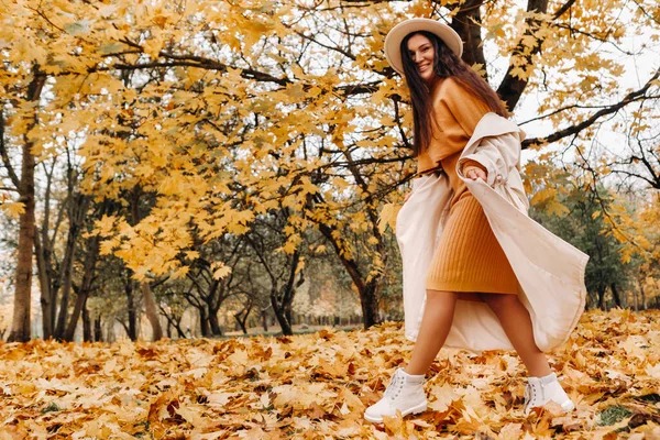 Ein fröhliches Mädchen in weißem Mantel und Hut lächelt in einem herbstlichen Park.Porträt einer lächelnden Frau im goldenen Herbst. — Stockfoto