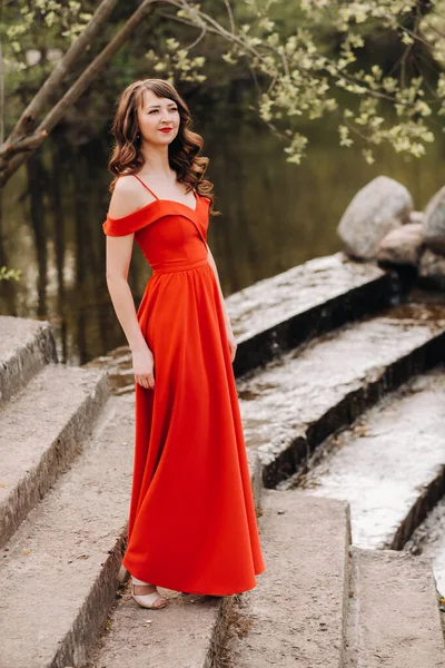 En ung vacker flicka med långt brunt hår, i en lång röd klänning kommer ner för trapporna nära sjön — Stockfoto