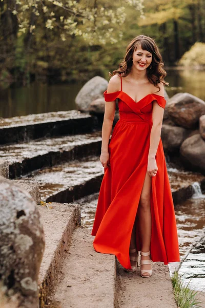 Ein junges schönes Mädchen mit langen braunen Haaren, in einem langen roten Kleid kommt die Treppe am See hinunter — Stockfoto