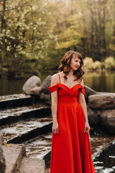 Uzun kahverengi saçlı, uzun kırmızı elbiseli güzel bir kız gölün yanındaki merdivenlerden aşağı iniyor. — Stok fotoğraf