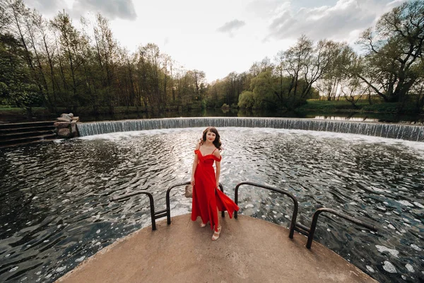 En ung vacker flicka med långt brunt hår, i en lång röd klänning kommer ner för trapporna nära sjön. — Stockfoto