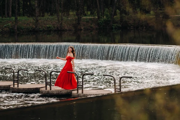 Portrait of a young beautiful laughing girl with long brown hair, in a long red dress in nature, near the lake, the time of year is spring — Stock Photo, Image