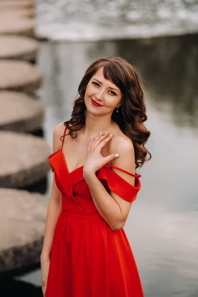 Mädchen im langen roten Kleid in der Nähe des Sees im Park bei Sonnenuntergang. — Stockfoto