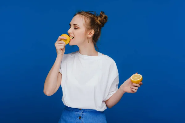 Una joven hermosa chica de pie sobre un fondo azul sosteniendo limones en su mano y mordiendo. — Foto de Stock