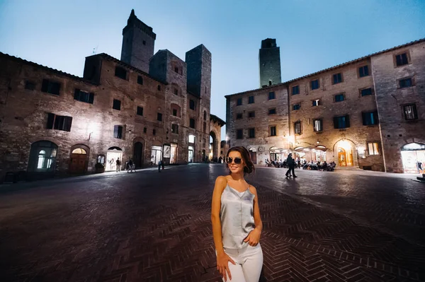 En flicka i byxor och en t-shirt med glasögon i nattstaden San Gimignano.En flicka går runt i staden i Italien.Toscana. — Stockfoto