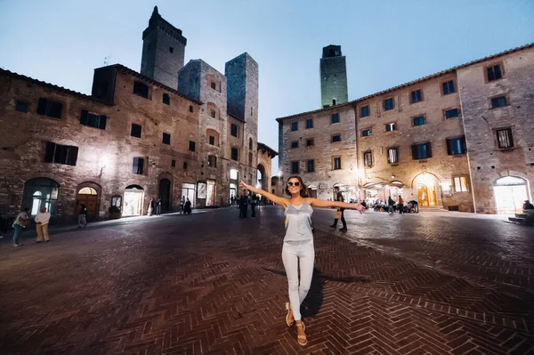 En flicka i byxor och en t-shirt med glasögon i nattstaden San Gimignano.En flicka går runt i staden i Italien.Toscana. — Stockfoto