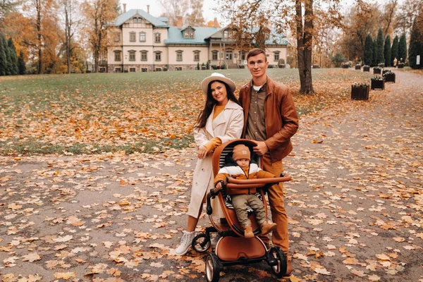 Padre y madre en un paseo con un niño en un cochecito en el parque de otoño en el fondo de la finca. Una familia pasea por el Parque Natural de Otoño Dorado — Foto de Stock