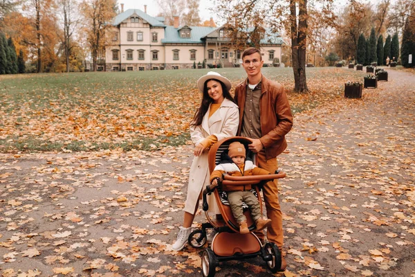 Far och mor på promenad med ett barn i en barnvagn i höstparken i bakgrunden av gården. En familj vandrar genom Gyllene höstens naturpark — Stockfoto