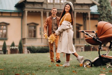 Anne ve baba, bir bebekle birlikte sonbahar parkında, arka planda bir bebek arabasında yürüyüşe çıktılar. Altın sonbahar doğa parkında bir aile yürüyor.