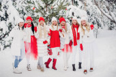 Velká skupina dívek s sklenicemi šampaňského v rukách stojí v zimním lese.Dívky v červených a bílých šatech s novoroční nápoji v zasněženém lese