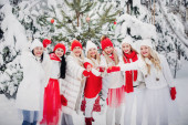 Velká skupina dívek s sklenicemi šampaňského v rukách stojí v zimním lese.Dívky v červených a bílých šatech s novoroční nápoji v zasněženém lese