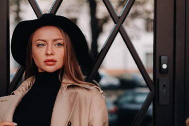 Şehir caddesinde siyah şapkalı, bej ceketli şık genç bir kadın. Kadın sokak modası. Sonbahar giysileri. Şehir tarzı..