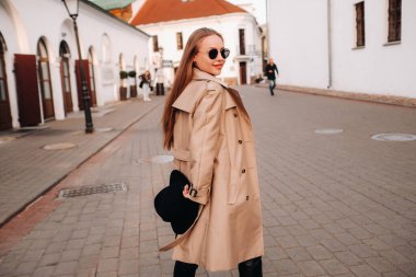 Bej ceketli, elinde siyah şapkalı ve gözlüklü şık genç bir kadın. Kadın sokak modası. Sonbahar giysileri. Şehir tarzı.