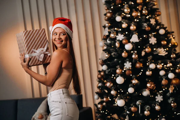 Una chica con un regalo de año nuevo en sus manos se levanta sobre el fondo de un árbol de Navidad en el interior —  Fotos de Stock