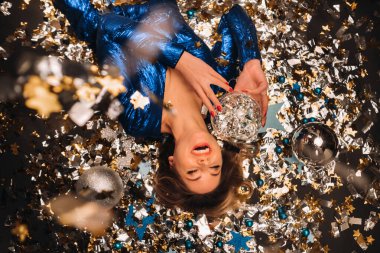 Mavi pullu elbiseli bir kadın yere düşmüş renkli konfetilerin altında gülümsüyor ve yatıyor..