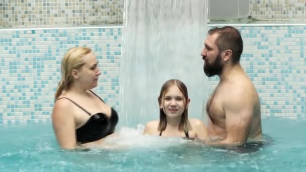 Una familia de tres bajo una cascada con un chorro de agua en una piscina azul. — Vídeos de Stock