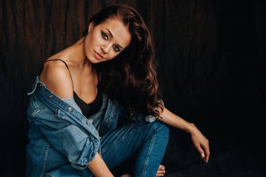 Uzun saçlı bir esmer, stüdyoda siyah bir arka planda, yerde oturuyor..