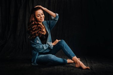 Uzun saçlı bir esmer, stüdyoda siyah bir arka planda, yerde oturuyor.