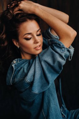 Uzun saçlı bir esmer, stüdyoda siyah bir arka planda, yerde oturuyor.