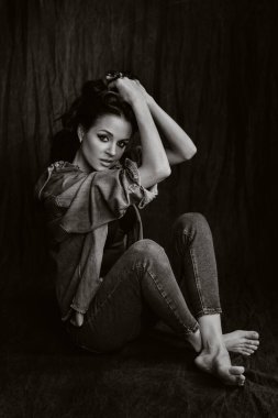 Stüdyoda uzun saçlı bir esmer, siyah arka planda yerde oturuyor. Siyah beyaz fotoğraf..