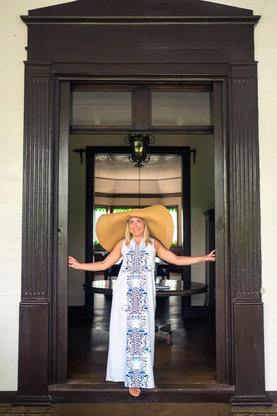 the woman in the big hat is smiling. a beautiful girl in a big hat and white dress smiles outside an old colonial building on the island of Mauritius.