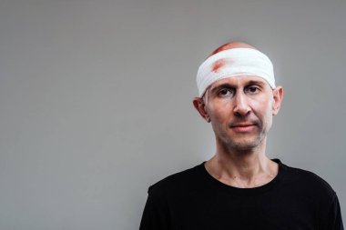 Gri arka planda siyah tişörtlü, başı bandajlı hırpalanmış bir adam duruyor.