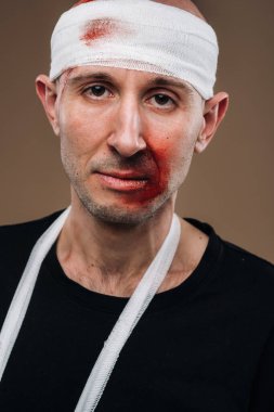 Kafası bandajlı ve kolunda alçı olan hırpalanmış bir adam gri bir arka planda duruyor.