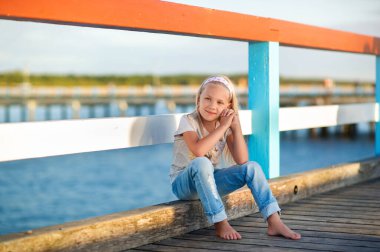 A little girl in jeans and a T-shirt sits on a pier near the Baltic Sea.Palanga, Lithuania