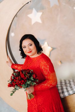 Kırmızı elbiseli bir kadın muhteşem bir stüdyoda duruyor ve elinde kırmızı güller ve çilekler tutuyor..