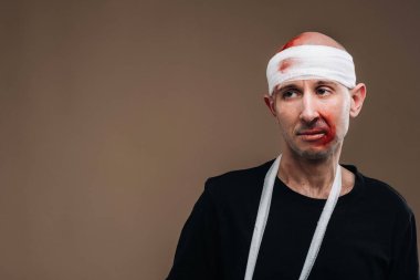 Kafası bandajlı ve kolunda alçı olan hırpalanmış bir adam gri bir arka planda duruyor..