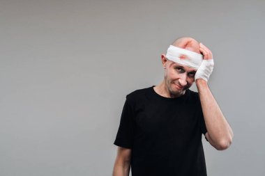 Siyah tişörtlü hırpalanmış ve hırpalanmış bir adam gri bir arka planda duruyor. Başı ağrıyan bir şekilde onu tutuyor..