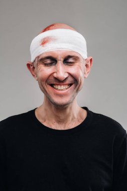 Gri arka planda siyah tişörtlü, başı bandajlı hırpalanmış bir adam duruyor..