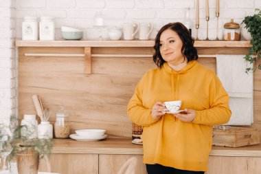 Turuncu kazaklı bir kadın evdeki mutfakta kahve içiyor..