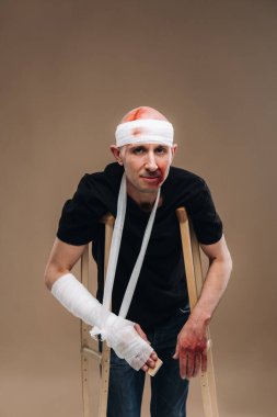 Kafası bandajlı ve kolunda alçı olan hırpalanmış bir adam gri bir arka planda koltuk değnekleriyle duruyor..