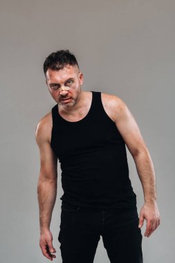 Siyah tişörtlü, uyuşturucu bağımlısı gibi görünen ve gri arka planda duran hırpalanmış bir adam..