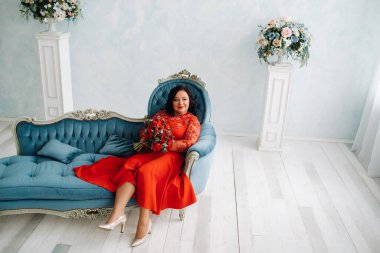 Kırmızı elbiseli bir kadın kanepeye oturur ve elinde bir buket kırmızı gül ve çilek tutar.