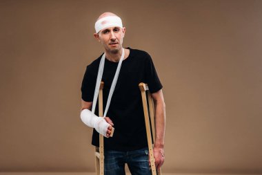 Kafası bandajlı ve kolunda alçı olan hırpalanmış bir adam gri bir arka planda koltuk değnekleriyle duruyor..