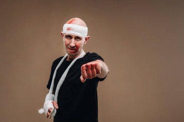 Kafası bandajlı ve kolunda alçı olan hırpalanmış bir adam gri bir arka planda duruyor..