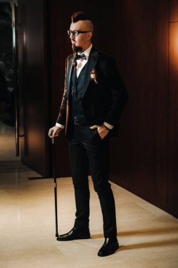 Smokinli ve papyonlu bir damat. Mohawk saç stili ve iç tarafında baston var..