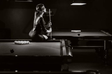 Gözü bağlı ve elinde ıstakayla bir bilardo kulübündeki masada oturan bir kız. Rus bilardosu. siyah beyaz fotoğraf.