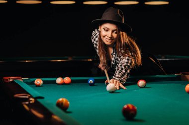 Bilardo kulübünde elinde ıstakayla şapkalı bir kız topa vuruyor. Bilardo oynuyor.