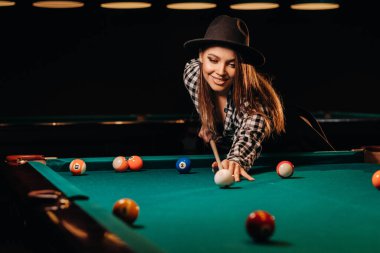A girl in a hat in a billiard club with a cue in her hands hits a ball.Playing billiards clipart