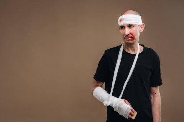 Kafası bandajlı ve kolunda alçı olan hırpalanmış bir adam gri bir arka planda koltuk değnekleriyle duruyor..